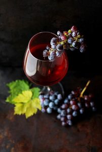 glass of red wine, bunches of purple grapes draped over glass and at bottom with leaves - Roberta Sorge on UnSplash