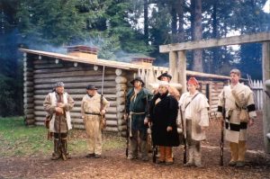 Fort Clatsop Park Rangers in brown period costumes standing in front of log building with green pine trees behind