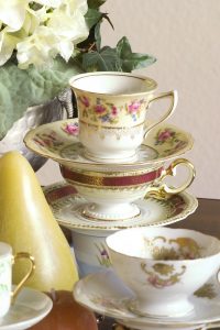 stack of three decorated fine china tea cups and saucers with gold napkin on left and white flowers in background