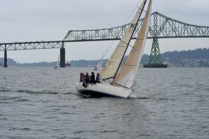Sailboat listing to starboard sailing on grey Columbia River toward Astoria-Megler bridge and Washington state coastline 