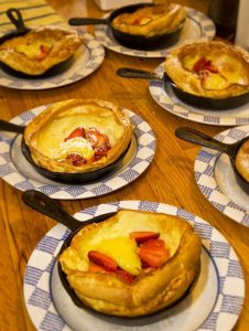 six individual black iron  skillets with Dutch pancakes and strawberries; skillets on blue & white check plates