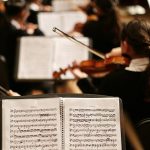 Sheet music on stand in foreground, more stands and backs of musicians playing violins in background by Adamara on UnSplash