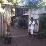 Fort Clatsop Park Ranger in white period costume standing by entrance to log fort - National Park Service