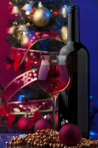 glass of red wine, dark wine bottle in front of glass cylinder of Christmas balls and ribbons, gold beads