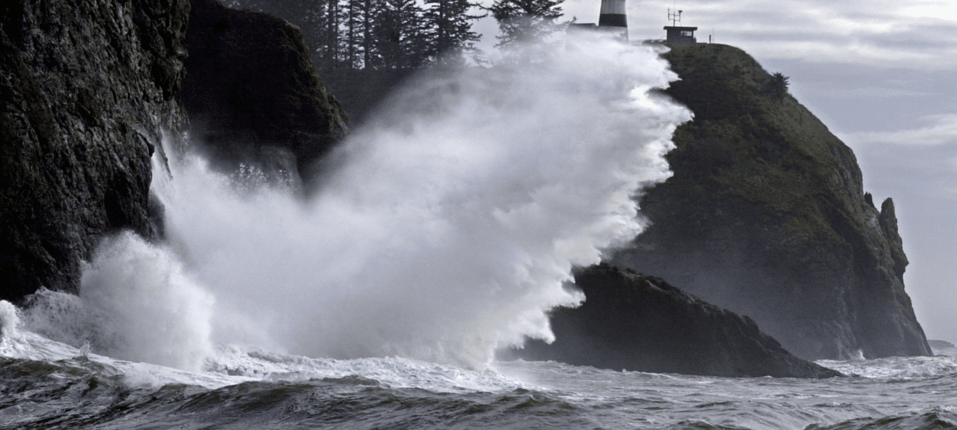 Powerful winter waves crashing against rocky cliffs near Astoria, Oregon, with a lighthouse overlooking the stormy Pacific Ocean.