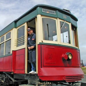 A bright red, yellow and green trolley by water with a conductor standing in doorway