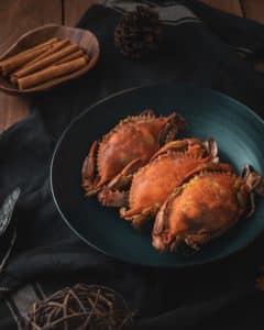 A plate of three crabs on a wooden table with cinnamon sticks and a pine cone nearby.