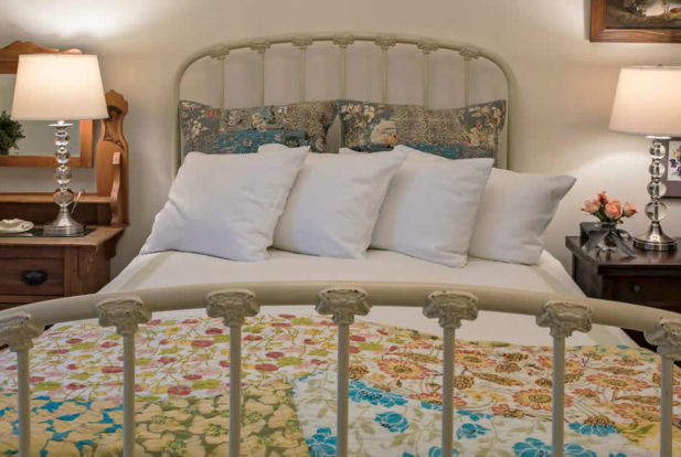 Painted metal bed made up in a bright quilt with fluffy pillows, a wooden nightstand and a wooden dresser.