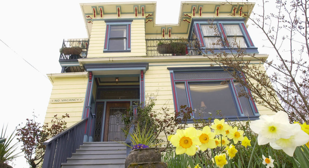 Yellow house with blue and burgundy trim and steps to the front door fronted by daffdils and trees.