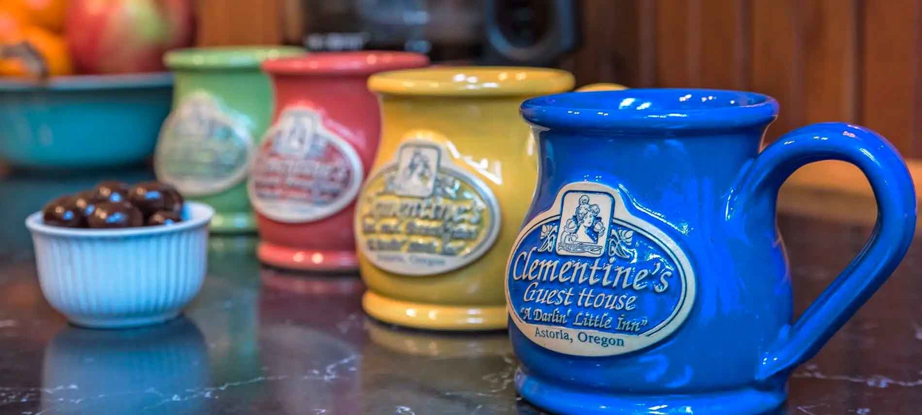 Row of brightly colored stoneware mugs with the Clementine's Guest House logo.