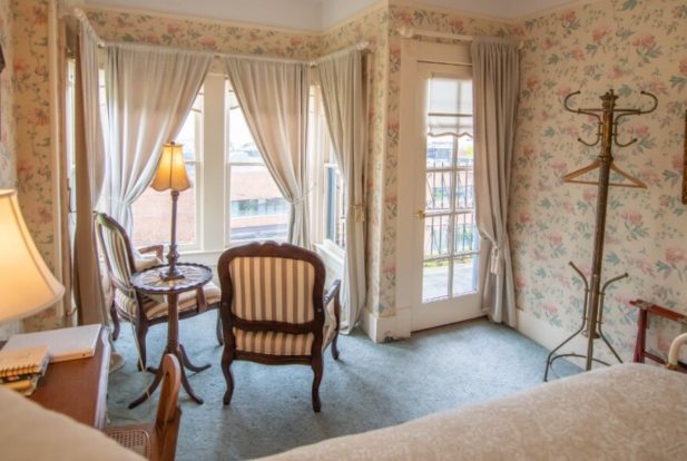Two brown chairs facing a large window with white drapes in a bedroom with flowered wallpaper.