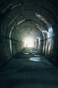 The silhouette of two people at the end of a dark stone tunnel.